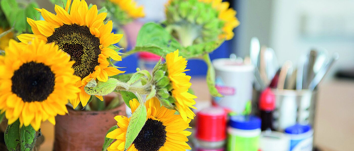 Vase mit Sonnenblumen steht auf einem Tisch. Auf einem Tablett stehen verschiedene Gewürze in farbigen Verpackungen.