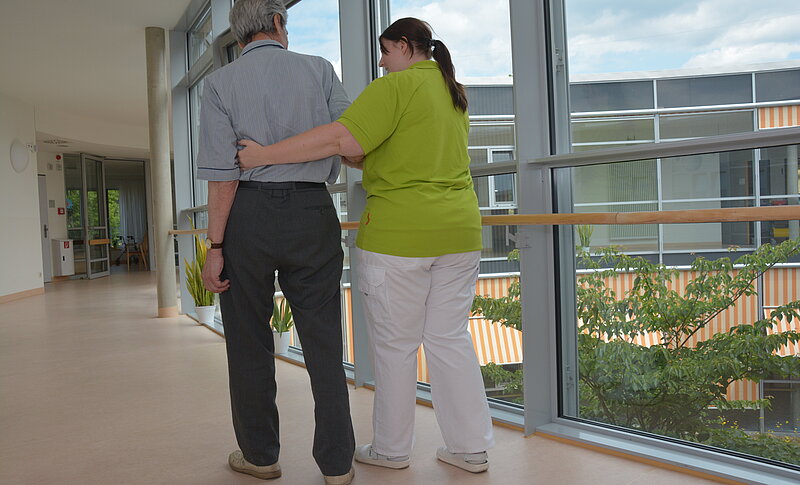 Eine Pflegefachkraft unterstützt einen älteren Herrn beim Gehen. Sie befinden sich im Flur der Klinik.