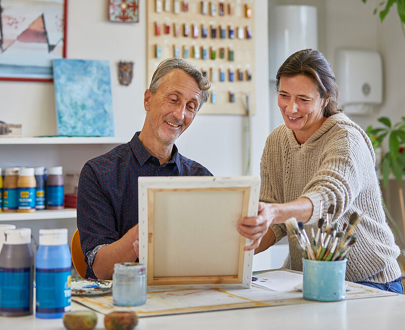 In einem Kreativraum unterhalten sich eine Frau und ein Mann über ein Kustnwerk. Im Hintergrund sind Farben, Bilder und Keramikstücke zu sehen.