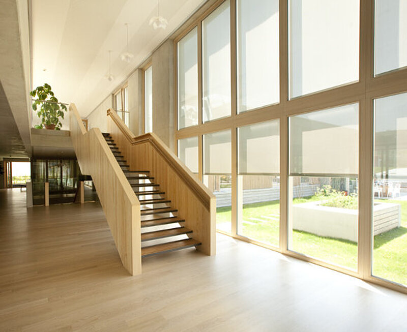 Erdgeschoss der Klinik mit Blick durch große Fensterflächen in den begrünten Innenhof. Eine Holztreppe führt in das nächste Geschoss.