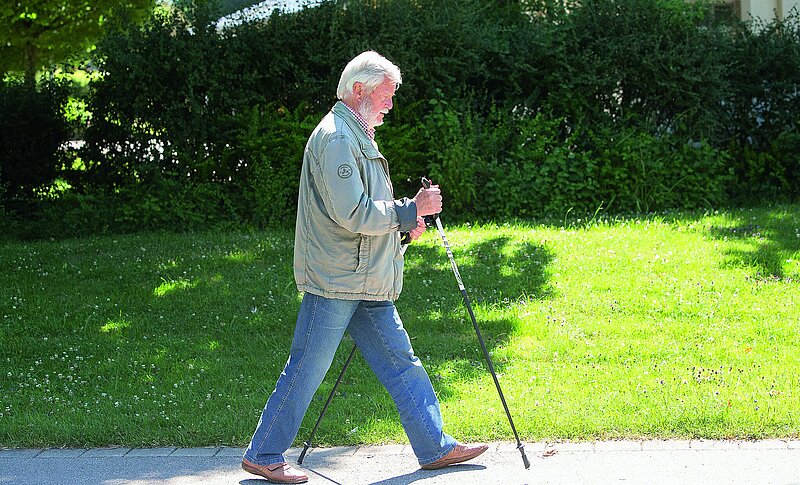 Ein älterer Mann geht mit seinen Walkingstöcken durch den Park. Es ist sonniges Wetter.