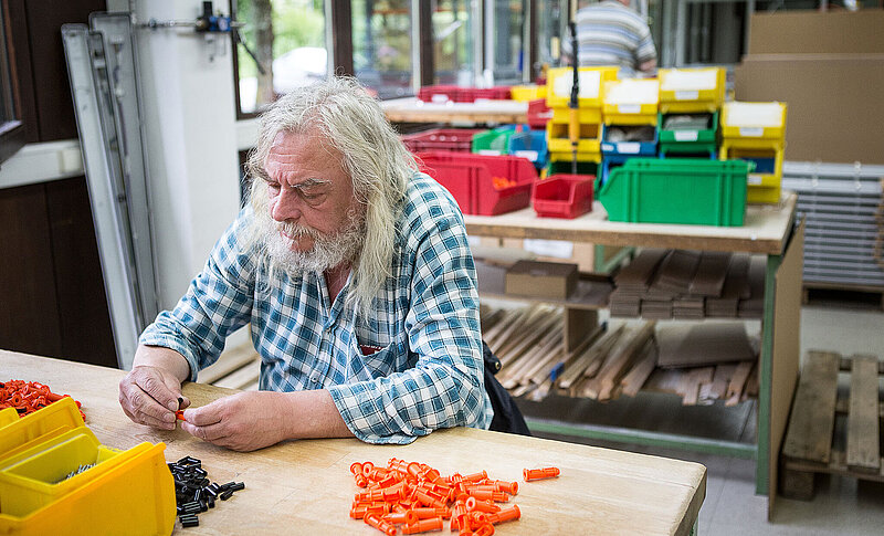 In einem Werkstattraum sitzt ein Mann mit langen Haaren und Bart am Tisch. Er setzt orange und schwarze Teile zusammen.